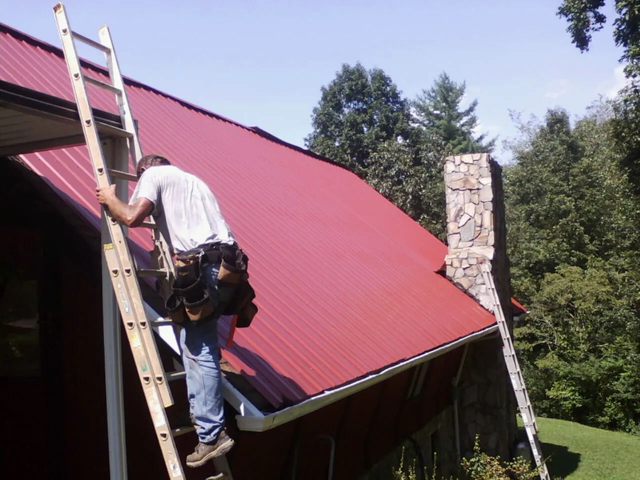 Roofer on a ladder beside a red metal roof and stone chimney — Soffit & Fascia Repair by Roofing Brothers in Ellisburg